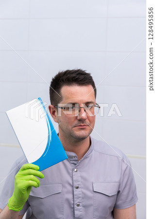 A Professional Male in Medical Gear Holding a Blue Document Folder in a Clean Clinical Environment with White Tiles 128495856