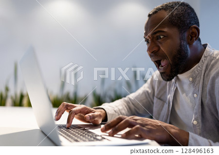Astonished male employee typing quickly on laptop and looking at screen with surprised expression 128496163