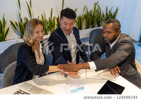Three diverse business colleagues sitting at table in modern office, stacking hands in unity 128496193
