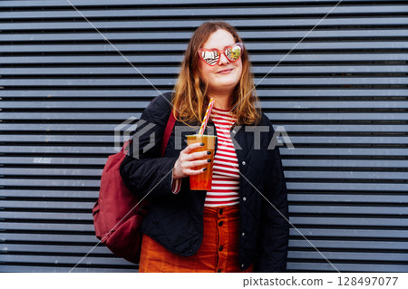Smiling plus size woman in heart shaped sunglasses and bright clothes drinking sugar flavored tapioca bubble tea near striped urban wall. Happy fashionable hipster overweight girl. Street fashion. 128497077