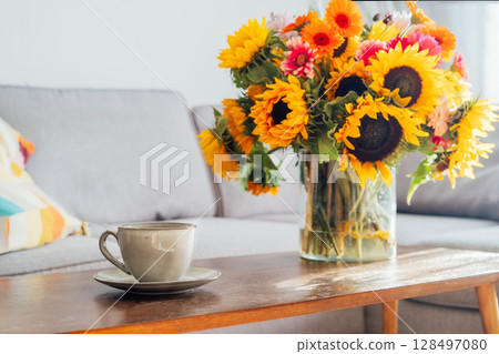 Focus on cup of hot drink and vase with huge sunflowers bouquet on wooden coffee table in modern cozy light living room with gray couch sofa. Cozy Design home decor. Summer mood, Relaxing rituals. Focus on cup of hot drink and vase with huge sunflowers bouquet on wooden coffee table in modern cozy light living room with gray couch sofa. Cozy Design home decor. Summer mood, Relaxing rituals. 128497080