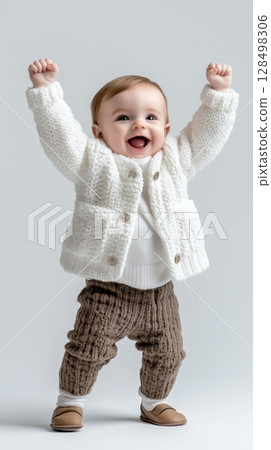 Cheerful two year old baby in a white jacket with raised arms, enjoying studio lighting 128498306