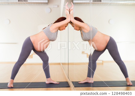 Pregnant woman is engaged in yoga. Exercise in a bent position, holding hands on a mirror wall 128498403