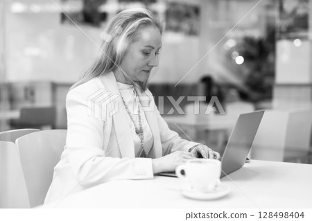 Business woman drinks coffee and works on laptop while sitting at table in cafe. View through window 128498404