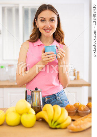 Smiling brown-haired woman enjoying cup of coffee in home kitchen Smiling brown-haired woman enjoying cup of coffee in home kitchen 128498430
