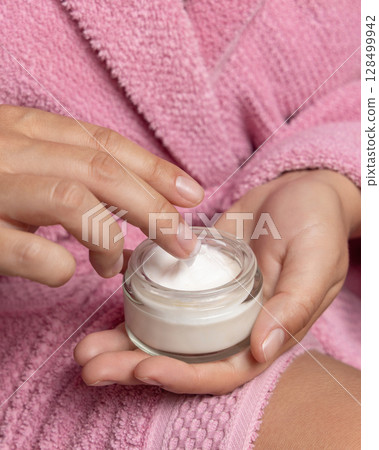 Young woman in pink bathrobe touching cream with finger and holding a cosmetic jar closeup 128499942
