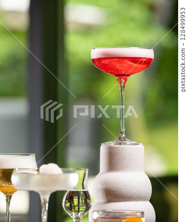 A vibrant red cocktail with a delicate white foam top is elegantly presented on a unique textured white stand, amidst other sophisticated drinks, against a lush green blurred background. A vibrant red cocktail with a delicate white foam top is elegantly presented on a unique textured white stand, amidst other sophisticated drinks, against a lush green blurred background. 128499953