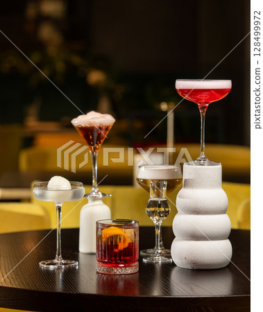 A richly colored red cocktail with a vibrant orange peel garnish is presented in a textured rocks glass, surrounded by other sophisticated drinks in a dimly lit bar setting. A richly colored red cocktail with a vibrant orange peel garnish is presented in a textured rocks glass, surrounded by other sophisticated drinks in a dimly lit bar setting. 128499972