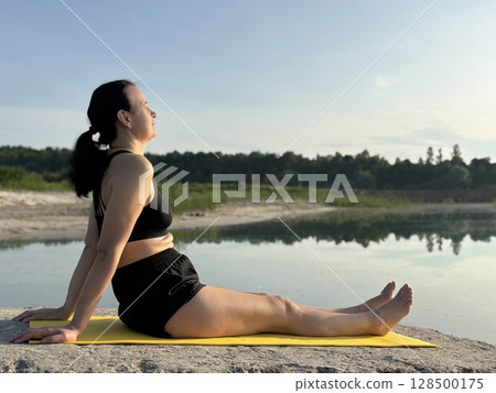 Adult woman sitting on yellow exercise mat and doing yoga outdoors by lake and forest. Summer fitness, sport activity in nature, healthy lifestyle, body care and mental balance 128500175