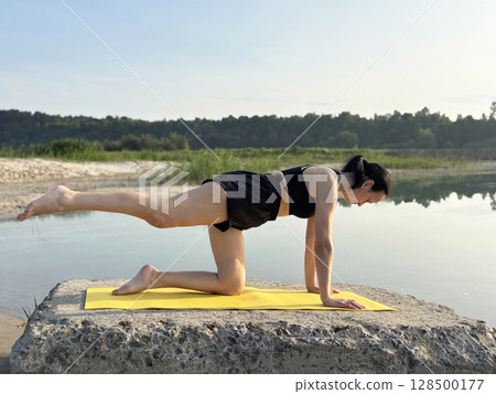 Adult woman doing yoga on yellow exercise mat outdoors by lake and forest. Summer fitness, sport activity in nature, healthy lifestyle, body care and mental balance Adult woman doing yoga on yellow exercise mat outdoors by lake and forest. Summer fitness, sport activity in nature, healthy lifestyle, body care and mental balance 128500177