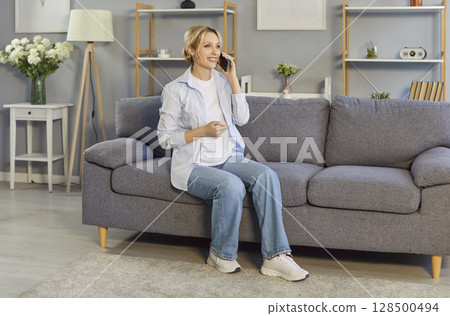 Happy young woman sitting on the sofa in the living room and talking on her mobile phone Happy young woman sitting on the sofa in the living room and talking on her mobile phone 128500494