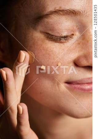 Soft glam. Macro view of smiling woman applying foundation dots on freckled cheek 128501435