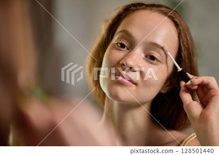 Young woman smiling while applying clear brow gel with natural expression and ease Young woman smiling while applying clear brow gel with natural expression and ease 128501440