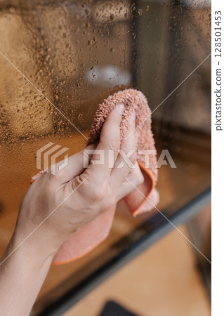 Vertical cropped shot of unrecognizable housewife wiping water droplets from window glass using orange microfiber cloth, professional cleaning worker performing window washing task, close-up. Vertical cropped shot of unrecognizable housewife wiping water droplets from window glass using orange microfiber cloth, professional cleaning worker performing window washing task, close-up. 128501453