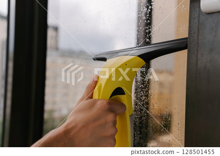Male hand using cordless vacuum cleaner for cleaning window, wiping away water drops on rainy day, ensuring spotless and hygienic indoor environment, close-up. Concept of household cleanliness. 128501455