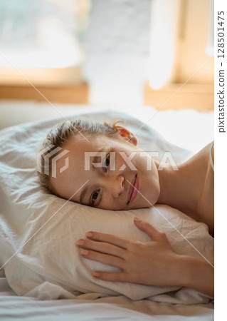 Peaceful young woman resting head on pillow with gentle smile in soft natural light Peaceful young woman resting head on pillow with gentle smile in soft natural light 128501475