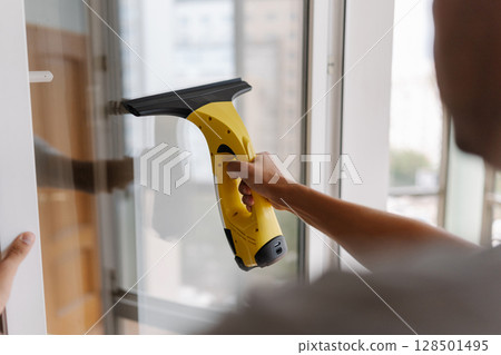 Close-up back view of cleaning service worker male using cordless vacuum cleaner to clean window, ensuring spotless and streak-free finish for pristine view, closeup. Concept of household chores. Close-up back view of cleaning service worker male using cordless vacuum cleaner to clean window, ensuring spotless and streak-free finish for pristine view, closeup. Concept of household chores. 128501495