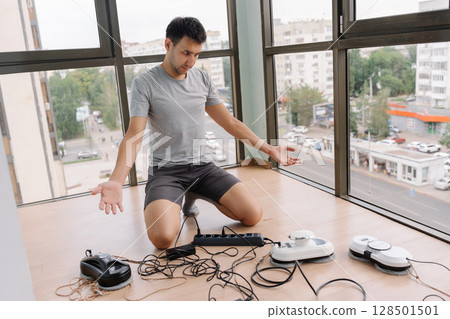 Portrait of shocked window cleaner male kneeling on floor and preparing multiple window cleaning robots for work, connecting them to power supply and showing palms to camera in home building 128501501