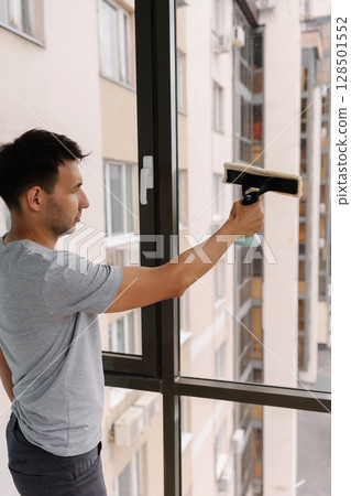 Vertical shot of young man spraying cleaning solution onto large window and wiping clean with squeegee, ensuring spotless and streak-free finish in building. Concept of cleanliness and housekeeping. Vertical shot of young man spraying cleaning solution onto large window and wiping clean with squeegee, ensuring spotless and streak-free finish in building. Concept of cleanliness and housekeeping. 128501552