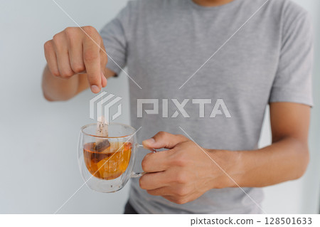 Close-up cropped shot of young unrecognizable man dipping tea bag into glass cup filled with steaming hot water, crafting soothing and aromatic beverage perfect for relaxation and enjoyment indoors. Close-up cropped shot of young unrecognizable man dipping tea bag into glass cup filled with steaming hot water, crafting soothing and aromatic beverage perfect for relaxation and enjoyment indoors. 128501633