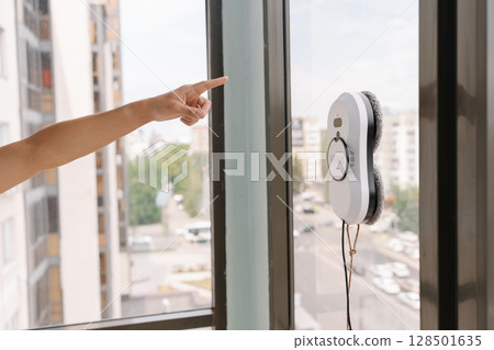 Homemaker gesturing toward robotic window cleaner working on tall glass building facade, representing advanced automated maintenance technology in modern workplace, close-up cropped shot. 128501635