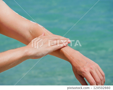 A man on the beach applies sunscreen lotion to his hands at the background of sea water. Skincare, sun protection and beach holidays concept A man on the beach applies sunscreen lotion to his hands at the background of sea water. Skincare, sun protection and beach holidays concept 128502660