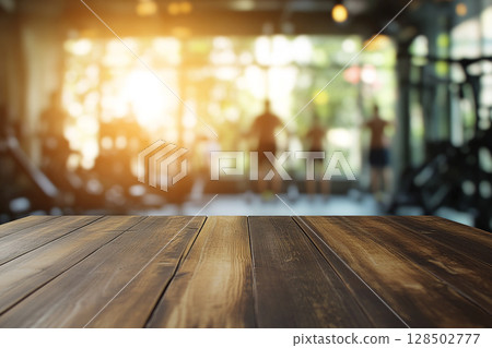 A close-up of a wooden table surface in the foreground, with a blurred gym interior and sunlit windows in the background. 128502777