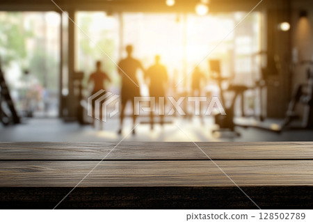 A close-up of a wooden table surface in the foreground, with a blurred gym interior and sunlit windows in the background. 128502789