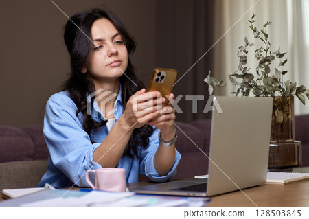 pretty woman in blue shirt holds smart phone uses laptop in home office 128503845