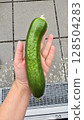 Fresh green cucumber lying on a human hand, outdoor setting with tiled floor and metal grate in background. Organic gardening harvest, homegrown vegetable. Natural light, top view. 128504283