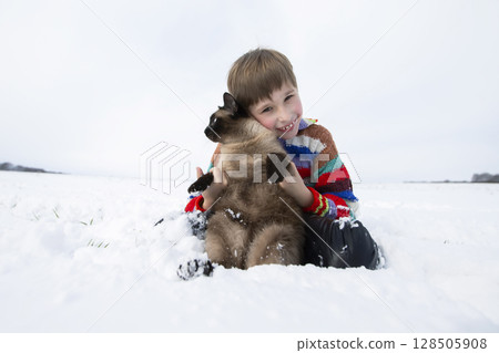 A little boy sits in the snow and holds a Siamese cat. 128505908
