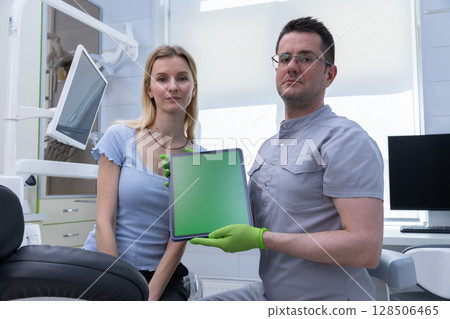 A confident dentist showcasing a digital tablet in a modern dental office alongside a patient, highlighting the use of technology in oral health care practices. 128506465