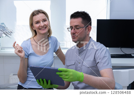 Patient Smiling with Dentist Discussing Treatment Options Using Tablet in Modern Dental Office Environment 128506488