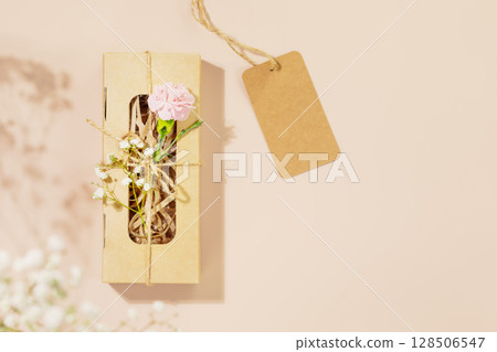 Romantic holiday composition with craft gift box with tender flowers and empty label on pink background. Eco friendly shopping, sale, packaging and delivery concept. Flat lay, copy space Romantic holiday composition with craft gift box with tender flowers and empty label on pink background. Eco friendly shopping, sale, packaging and delivery concept. Flat lay, copy space 128506547