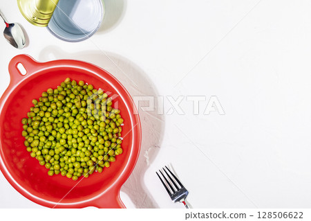 Green canned peas in a red plastic colander next to an open tin can, fork and spoon on white table. Canned and conservation food. Flat lay, copy space Green canned peas in a red plastic colander next to an open tin can, fork and spoon on white table. Canned and conservation food. Flat lay, copy space 128506622