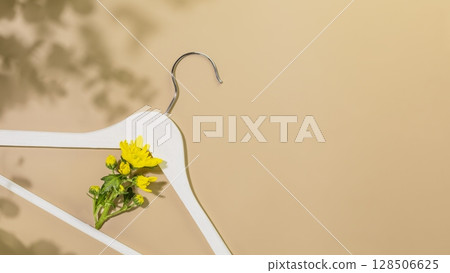 Eco friendly still life with white wooden hanger with yellow flower on beige background with plant shadows. Sustainable shopping, buying or sales concept. Flat lay, copy space Eco friendly still life with white wooden hanger with yellow flower on beige background with plant shadows. Sustainable shopping, buying or sales concept. Flat lay, copy space 128506625
