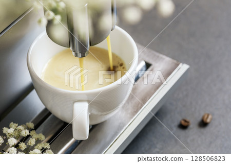 Coffee machine makes espresso. Professional coffee brewing. Cup of fresh hot espresso with coffee beans on grey natural background with flower. Coffee break. Romantic morning. Close up, soft focus 128506823