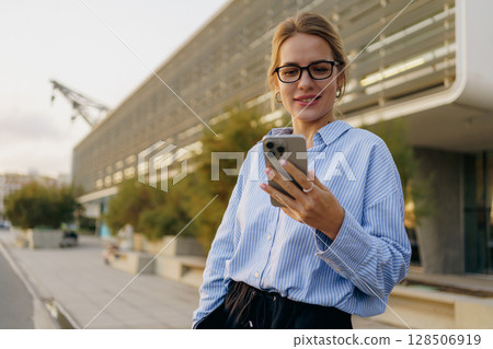 A Young Woman is Using a Smartphone While Enjoying the Outdoors in a Modern Urban Setting 128506919