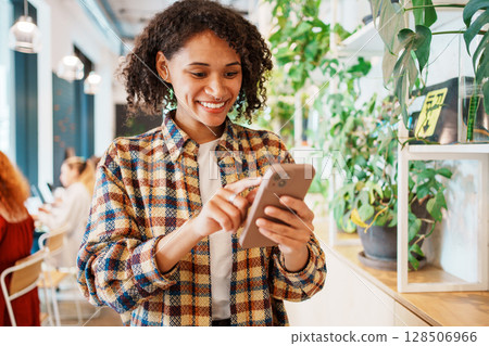 A cheerful woman happily engaging with her smartphone while enjoying her time in a cozy cafe 128506966