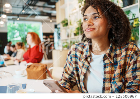 A young woman sits in a trendy cafe, using her smartphone and engaging with her surroundings 128506967