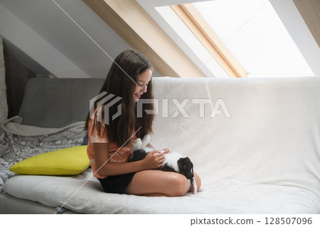 Smiling girl playing with black and white rabbit indoors. 128507096