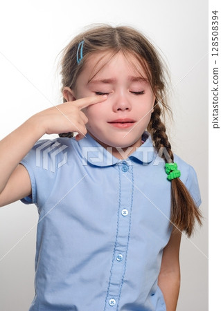 Young girl crying, wiping away tears with her hand. Sad expression, light blue shirt. 128508394