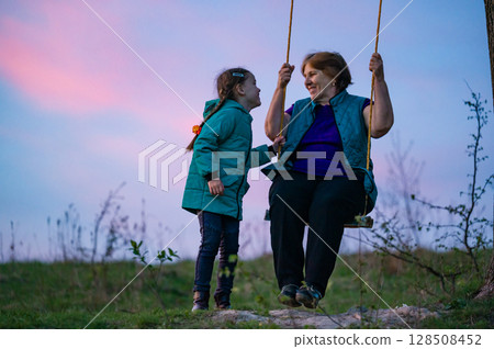 Grandmother and granddaughter sharing a joyful swing ride at sunset. A heartwarming moment of intergenerational connection. 128508452