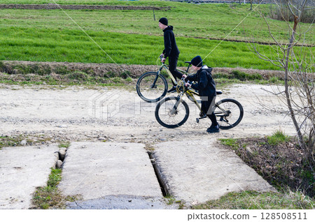 Two boys on mountain bikes ride along a rural road, navigating a damaged section of pavement. A sunny day in the countryside. Two boys on mountain bikes ride along a rural road, navigating a damaged section of pavement. A sunny day in the countryside. 128508511