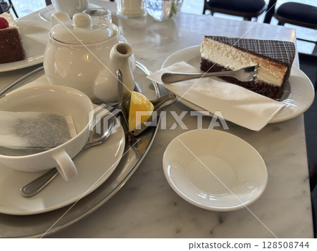 Piece of chocolate cake on white plate with tea bag in cup and tea-pot, lemon near it, sweet dessert in cafe, side view close up, selective focus 128508744