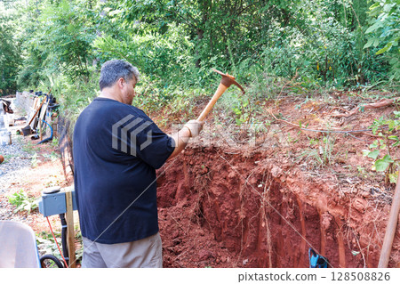Worker is actively digging into red soil in forested area with digging tool. Worker is actively digging into red soil in forested area with digging tool. 128508826