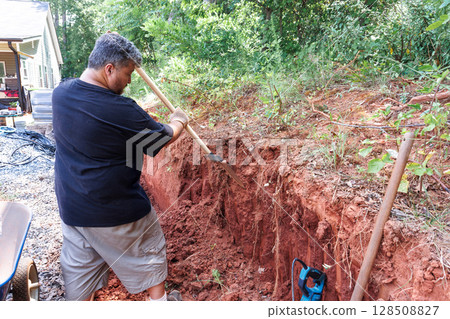 Work man works diligently to dig trench in red clay near house, surrounded by greenery tools 128508827