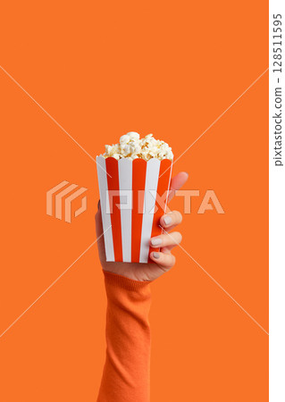 Womans hand holding striped box with popcorn on an orange background 128511595