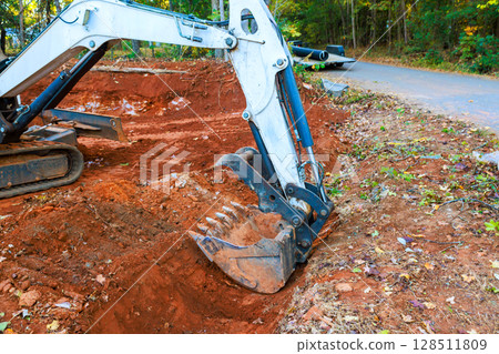 Heavy machinery is digging trench in red dirt near gravel road surrounded by trees under works daytime operations Heavy machinery is digging trench in red dirt near gravel road surrounded by trees under works daytime operations 128511809