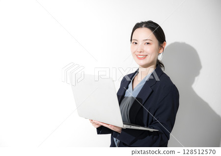 Business image of a young cute woman standing in a suit holding a laptop and looking at the camera, upper body Business image of a young cute woman standing in a suit holding a laptop and looking at the camera, upper body 128512570